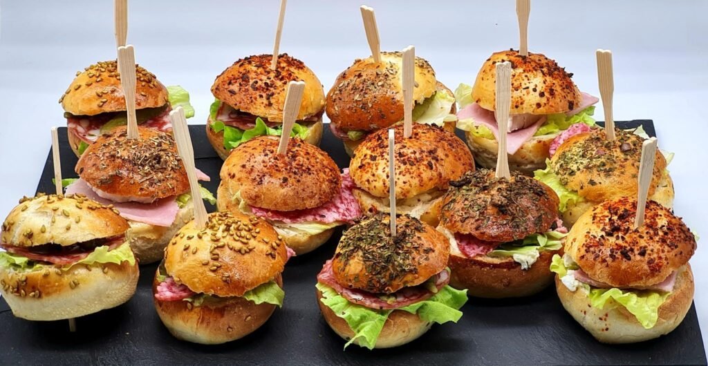 Mini slider sandwiches with ham, salami, lettuce and cheese on a black slate, each with a wooden skewer