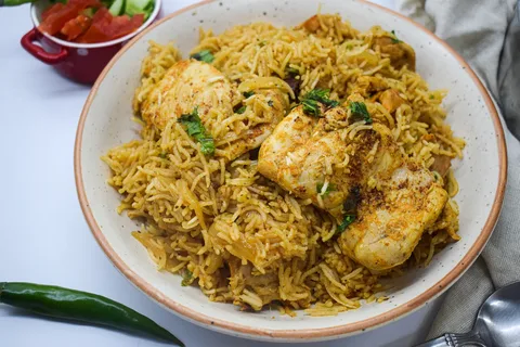 Chicken biryani with golden spiced rice and chicken pieces in a white bowl, with tomato salad in the background