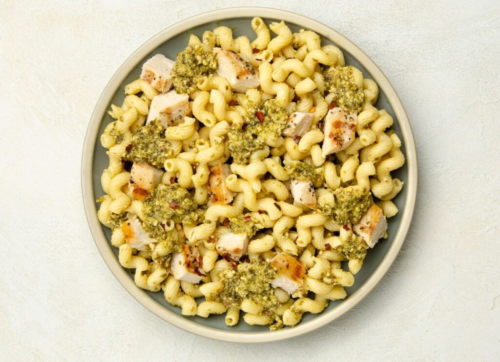 Overhead view of cavatappi pasta with pesto sauce, grilled chicken pieces, and red pepper flakes on a plate