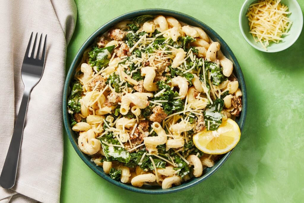 Overhead view of cavatappi pasta with kale, sausage, and Parmesan cheese in a blue bowl, with lemon wedge, fork, and side of cheese